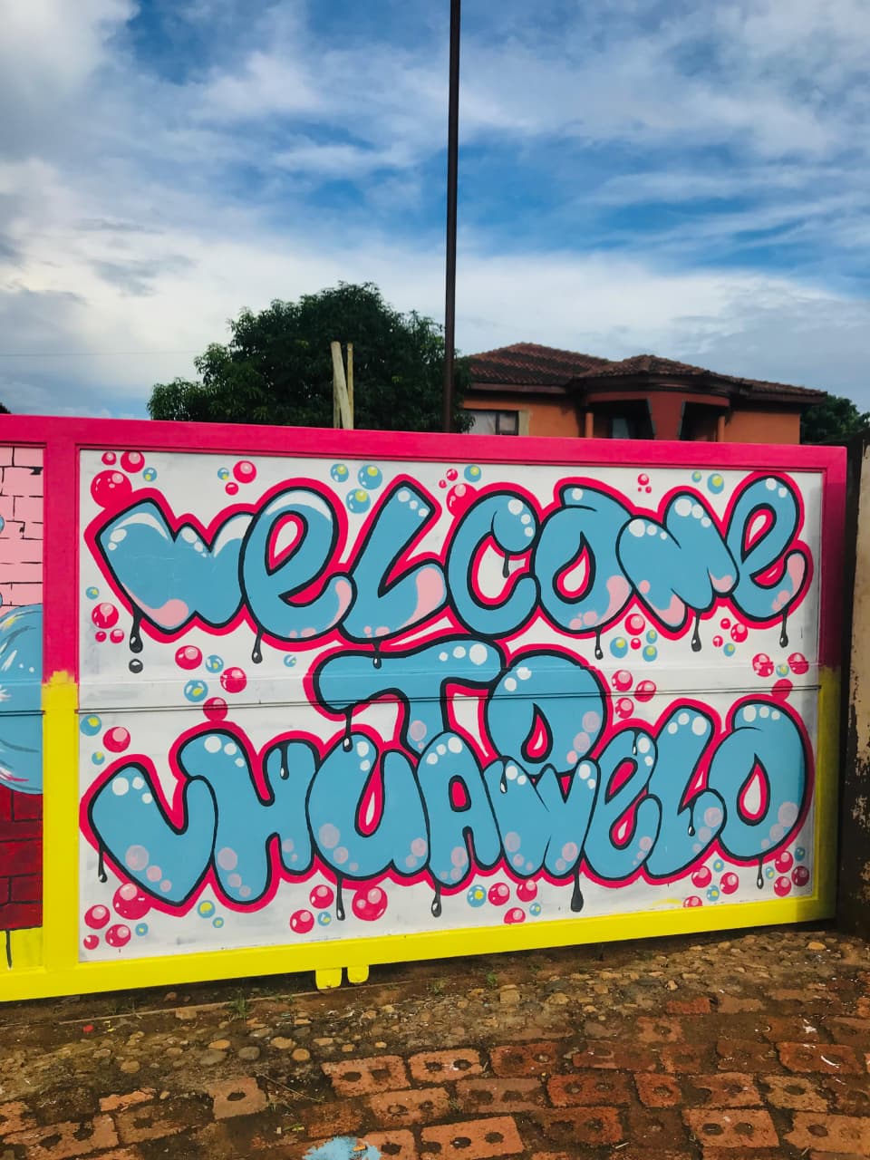 Sliding gate painted with the words Welcome to Vhuawelo in blue bubble letters with a pink frame