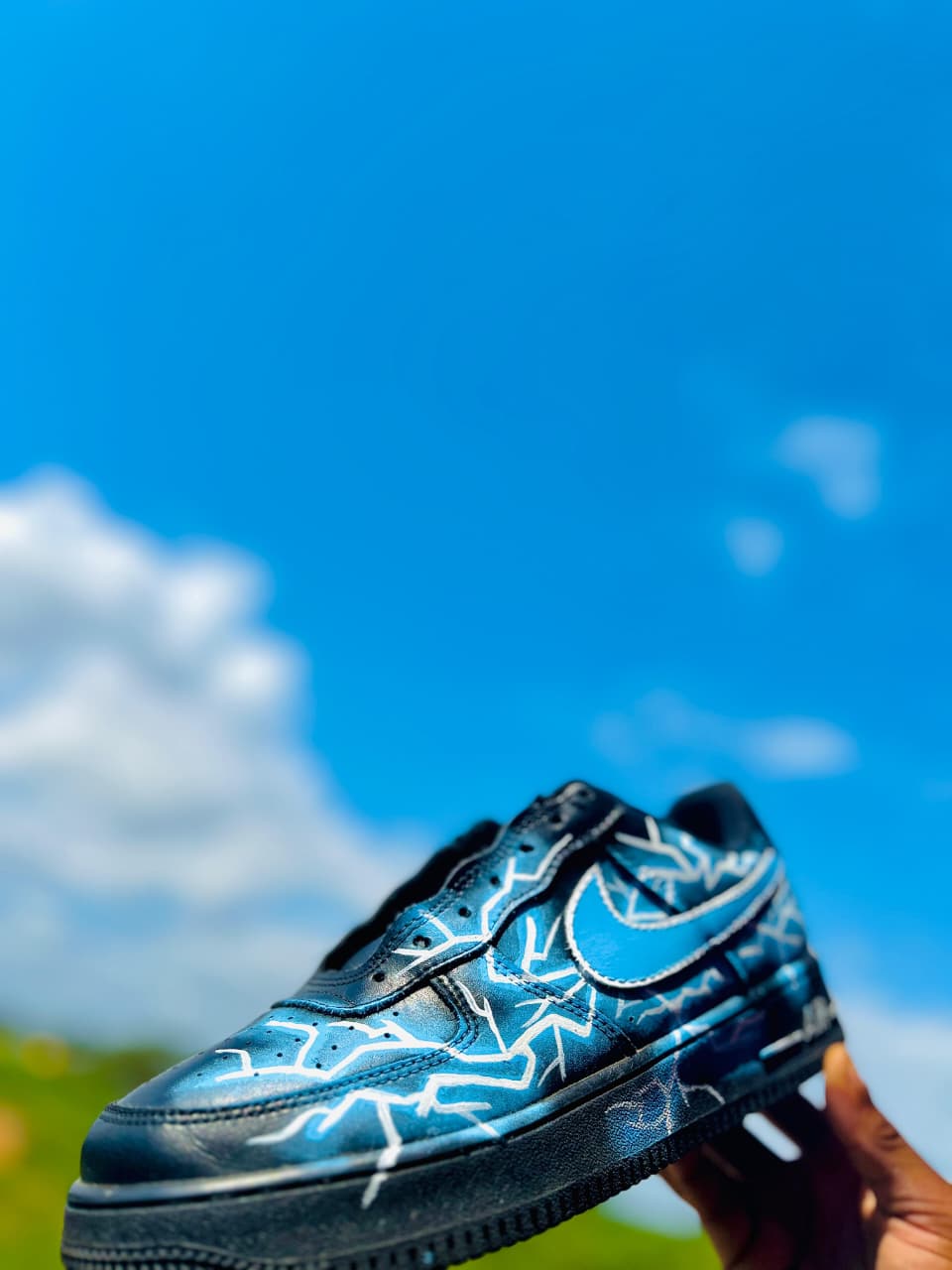 Hand-painted black Nike Air Force 1 with an electric-blue fade and white lightning lines, held up against a clear blue sky