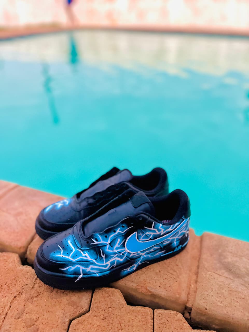A pair of hand-painted black sneakers with blue lightning artwork resting on the edge of a swimming pool